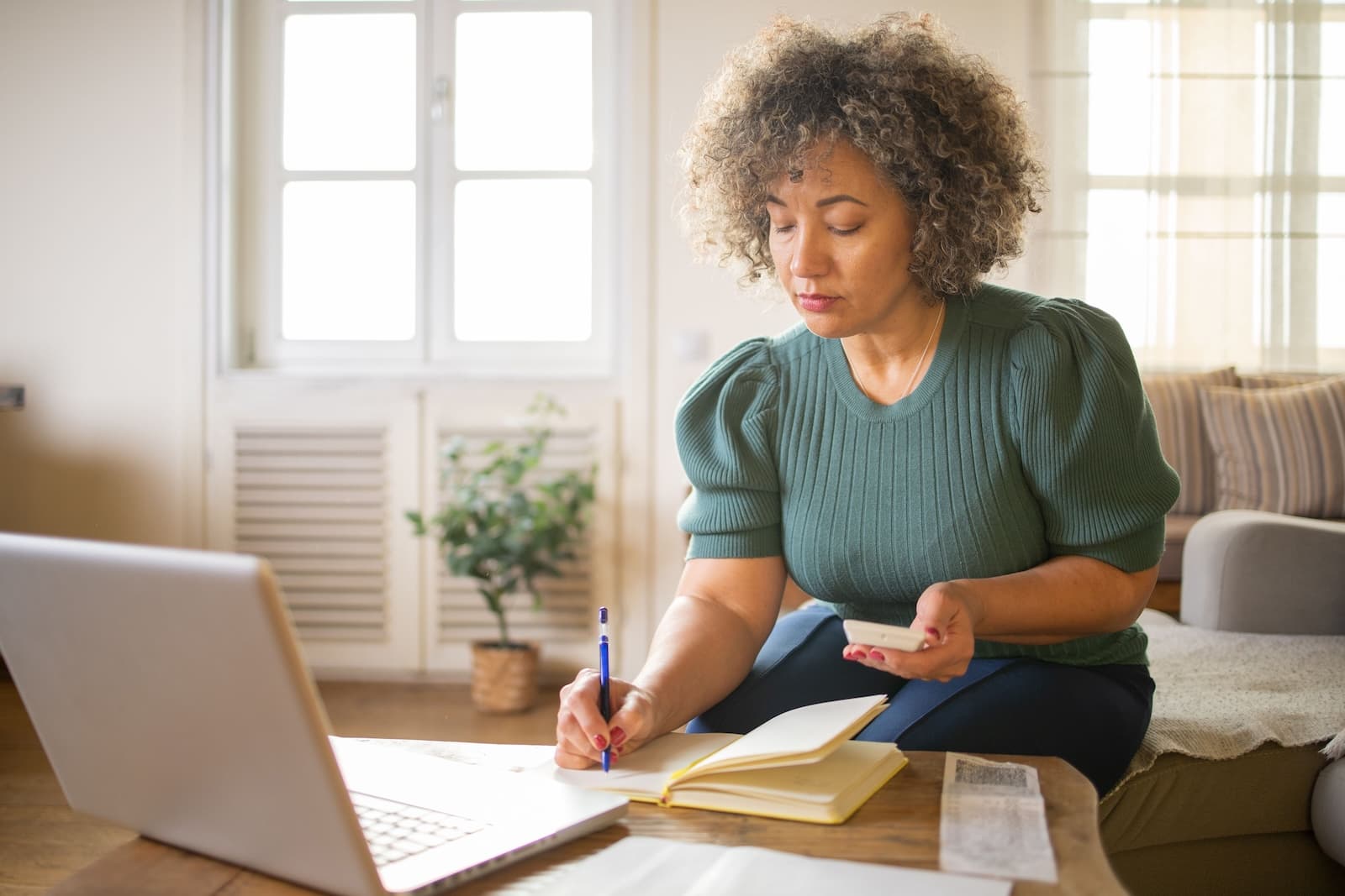 A woman writing in notebook with calculator in hand and laptop in front